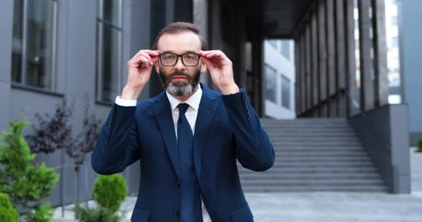 Portret białego mężczyzny w średnim wieku stojącego na zewnątrz, dotykającego i naprawiającego okulary i patrzącego w kamerę z uśmiechem. Starszy mężczyzna biznesmen w garniturze i krawacie z wesołą twarzą w centrum miasta. — Wideo stockowe