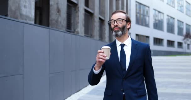 Кавказский красивый элегантный бизнесмен в костюме, галстуке и очках весело гуляет по утрам на работу и пьет кофе на вынос. Счастливый мужчина средних лет потягивает напитки и гуляет по городу. — стоковое видео