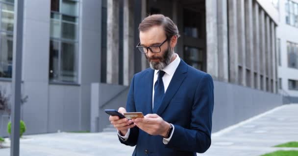 Biały, przystojny biznesmen w garniturze, krawacie i okularach stukający smartfonem i robiący zakupy online kartą kredytową. Szczęśliwy mężczyzna w średnim wieku kupuje i płaci w Internecie przez telefon komórkowy na ulicy miejskiej. — Wideo stockowe