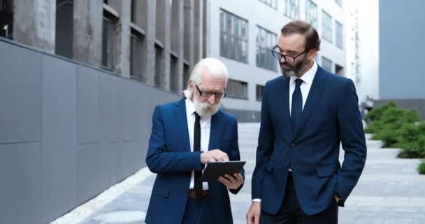 Viejo hombre caucásico jefe caminando en la calle de la ciudad con el socio de negocios masculino y mostrando información sobre el dispositivo de la tableta. Empresarios hablando y discutiendo sobre el contorno al aire libre. Trabajo y paseos. — Vídeos de Stock