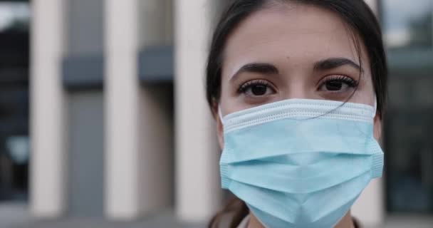 Close-up van vrolijke blanke jonge donkerharige vrouw die in de buitenlucht staat met een medisch masker op haar gezicht en naar de camera kijkt. Gelukkige mooie vrouw alleen op straat in quarantaine. Gezondheidsconcept — Stockvideo