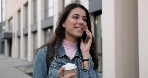 Közelkép portré vidám fiatal kaukázusi nő sétál az utcán a kávé-to-go és csevegés okostelefonon. Gyönyörű örömteli nő beszél mobilon a szabadban és mosolyog. Beszélgetés — Stock videók