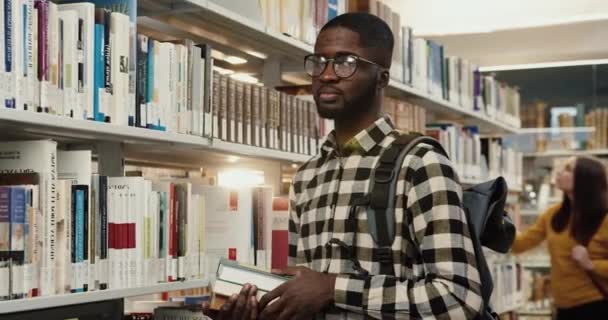 Портрет молодого африканского успешного студента, стоящего рядом с книжными полками в библиотеке и улыбающегося, глядя в камеру. — стоковое видео