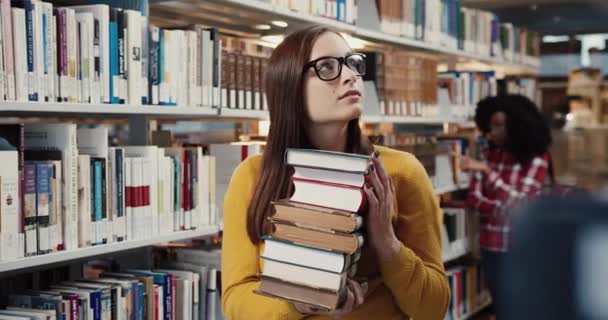 Портрет молодого успішного студента, який стоїть в бібліотеці, тримає велику стопку книг і посміхається дивитися на камеру . — стокове відео