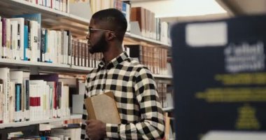 Kütüphanede kitaplıkların yanında duran, elinde kitap tutan ve kameraya bakıp gülümseyen Afro-Amerikan öğrencinin portresi..