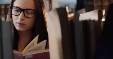 Kütüphanede kitap raflarının arasında duran ve dikkatle kitap okuyan gözlüklü güzel beyaz kadın. .
