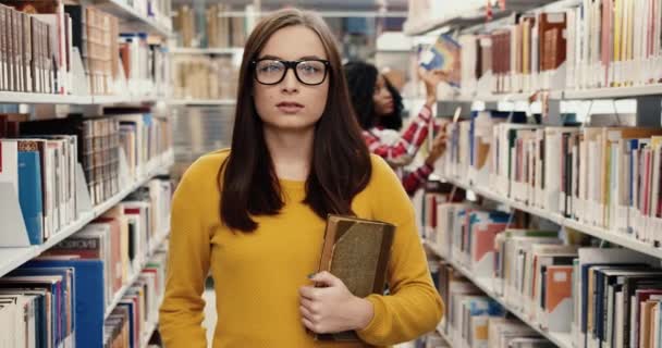 Retrato de jovem estudante nerd feliz feminino em pé na biblioteca segurando livro e sorrindo olhando para a câmera. — Vídeo de Stock