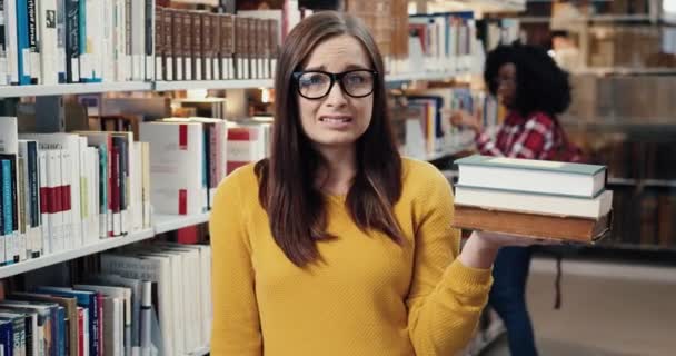 Крупним планом портрет молодої студентки-жінки в окулярах, що стоять в бібліотеці і розчаровані поглядом на стек книг для читання для лекцій . — стокове відео