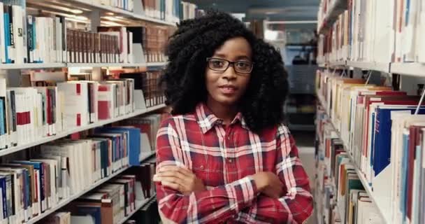 Porträtt av ung glad afrikansk amerikansk kvinnlig student med lockigt hår i glasögon står bland bokhyllor i biblioteket och ler tittar på kameran. — Stockvideo