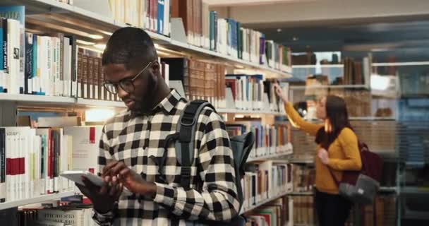 Mladý úspěšný student stojí v knihovně poblíž knihoven a píše na svém tabletu. Portrét chytrého afrického amerického studenta. — Stock video