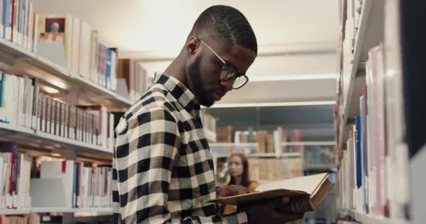 Młody Afroamerykanin w okularach stojący w bibliotece uniwersyteckiej i trzymający starą książkę przewracając kartki. — Wideo stockowe