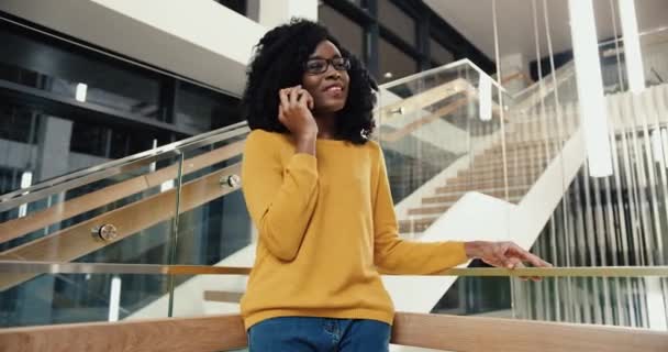 Joven hermosa africana americana estudiante en glasess de pie en el vestíbulo del campus y hablando con amigos por teléfono. — Vídeo de stock