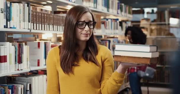 Portret van ongelukkige student staat in de bibliotheek en onderzoekt angstig het aantal boeken dat snel gelezen moet worden. — Stockvideo