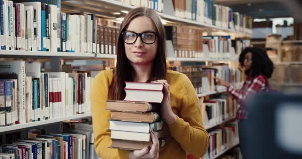 Potret siswa perempuan cantik dengan kacamata berdiri di perpustakaan memegang banyak buku dan menikmati belajar tersenyum melihat kamera. — Stok Video