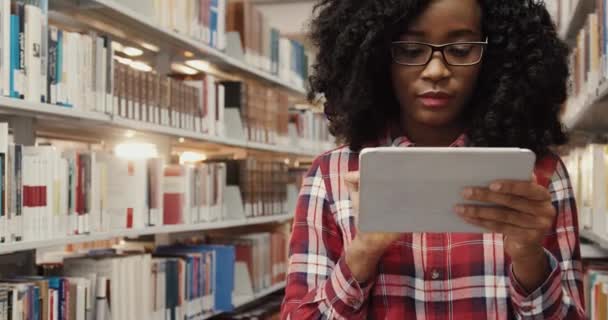 Κοντινό πλάνο της νεαρής όμορφης αφροαμερικάνας φοιτήτριας με γυαλιά που στέκεται ανάμεσα σε σειρές βιβλίων στη βιβλιοθήκη και ψάχνει για πληροφορίες στο tablet. — Αρχείο Βίντεο