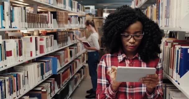 Умная красивая студентка в очках, стоящая среди книжных полок в библиотеке и ищущая информацию в планшете, смотрит вверх и улыбается. — стоковое видео