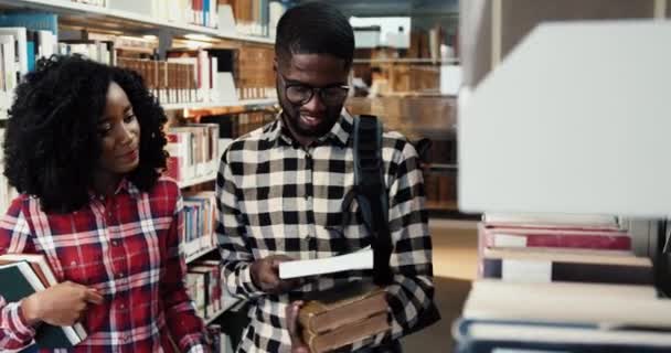 Young couple of African American students are looking for literature in library to study. Smart guy and girl looking book standing near bookshelves.