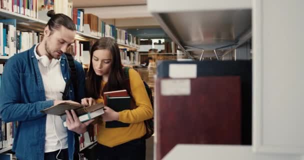 Νεαρό έξυπνο κορίτσι και όμορφος άντρας που στέκεται στη βιβλιοθήκη ερευνώντας βιβλία και συζητώντας κείμενα. Δύο φίλοι μαθητές προετοιμάζονται για τις εξετάσεις. — Αρχείο Βίντεο