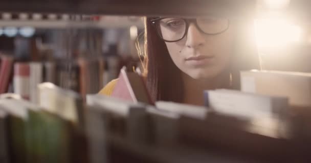 Крупным планом молодой красивой умной кавказки, стоящей в библиотеке среди полок и пристально ищущей книги. — стоковое видео
