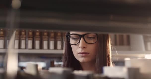 Close up of young beautiful smart caucasian girl in glasses standing in library among shelves and intently reading book. — Stock Video