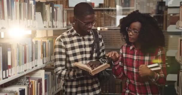 Unga afroamerikanska par smarta studenter går genom biblioteket bland hyllor och diskuterar bokhållare i sina armar. — Stockvideo