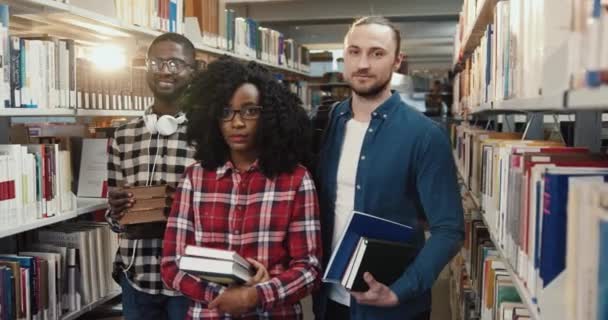 Porträtt av tre smarta multinationella studenter som står i biblioteket och håller böcker. Bästa vänner förbereder sig för tentor. — Stockvideo