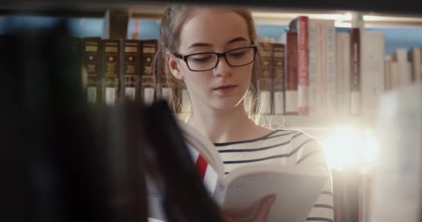 Крупним планом молода красива розумна кавказька дівчина стоїть в бібліотеці серед полиць і свідомо читає книгу . — стокове відео