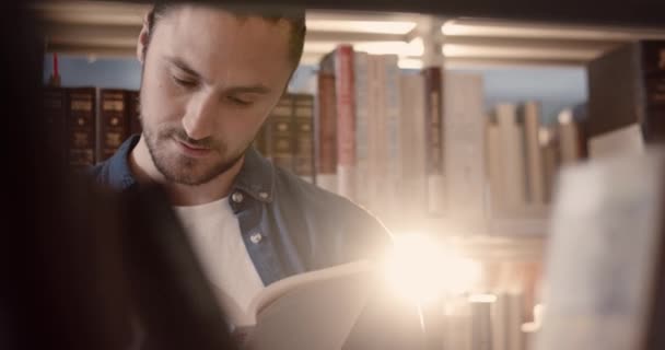 Close-up van jong knap slim Kaukasisch guy staan in de bibliotheek tussen planken en aandachtig lezen interessant boek. — Stockvideo