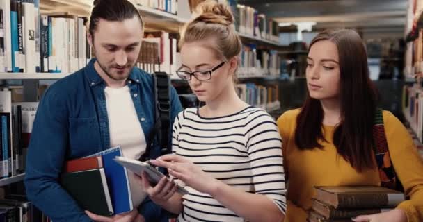 Крупный план портрета трех счастливых белых умных друзей, стоящих в библиотеке и держащих книги и планшеты, смотрящих в камеру, сморщиваясь. — стоковое видео