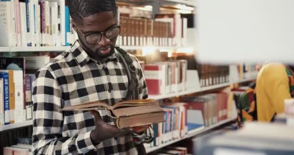 Close-up van de jonge Afro-Amerikaanse student met een bril in de universiteitsbibliotheek en leesboek, bladerend door de pagina 's. — Stockvideo