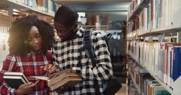 Close up de jovens afro-americanos casal de estudantes em pé na biblioteca e rever livro interessante e discutir o seu conteúdo. — Vídeo de Stock