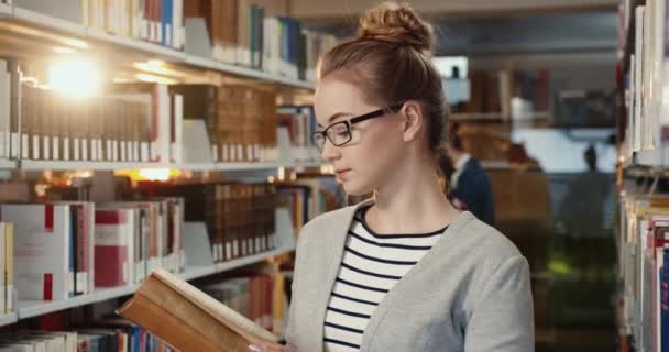 Close-up van jong slim student meisje met bril staan in de leeszaal tussen planken kijken naar camera glimlachen. — Stockvideo