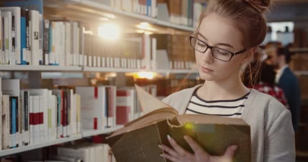 Primo piano della giovane studentessa intelligente con gli occhiali in piedi nella sala di lettura tra gli scaffali e libro di lettura intensamente. — Video Stock