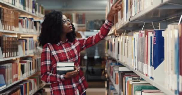 年轻美丽的非洲裔美国女孩，留着卷发，戴着眼镜，走在图书馆的一排中寻找必要的书籍. — 图库视频影像