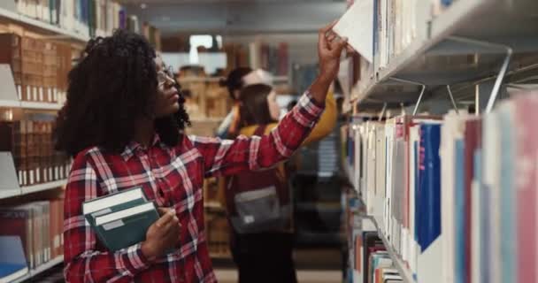 Νεαρή έξυπνη Αφρο-Αμερικανίδα με σγουρά μαλλιά σε γυαλιά περπατά στις σειρές της βιβλιοθήκης αναζητώντας λογοτεχνία για διάλεξη. — Αρχείο Βίντεο