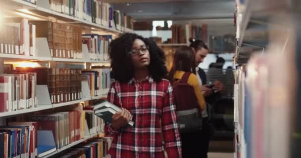 Mladá Afroameričanka s kudrnatými vlasy v brýlích prochází knihovnou řádky v hledání literatury pro volný čas. — Stock video