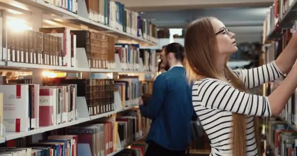 Молодая умная кавказская блондинка в очках стоит среди книжных полок и выбирает литературу для свободного чтения. — стоковое видео