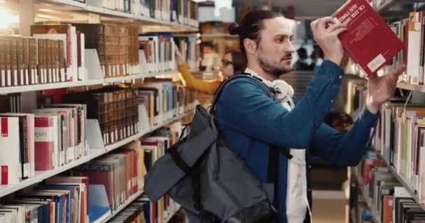 Jonge knappe blanke student met witte koptelefoon staat in de bibliotheek tussen de rijen op zoek naar het nodige boek. — Stockvideo