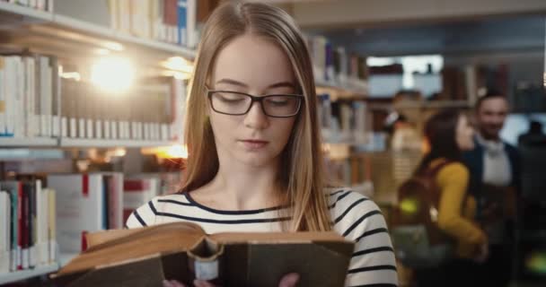 Zbliżenie się młody inteligentny blondynka student w okulary stojąc w bibliotece i uważnie studiuje książkę. — Wideo stockowe