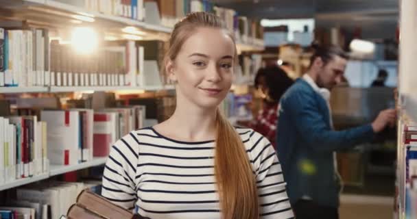 Närbild porträtt av ung blond kvinna student stående i biblioteket håller böcker och tittar på kameran ler. — Stockvideo