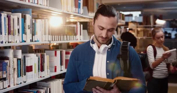 Retrato de un joven estudiante en camisa de mezclilla con auriculares de pie en la biblioteca lee libro mira hacia arriba y sonríe a la cámara. — Vídeo de stock