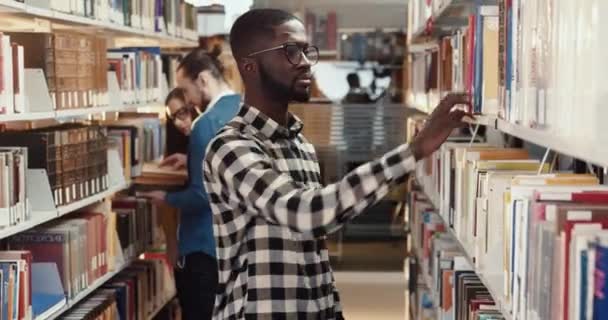 Un joven afroamericano con camisa a cuadros con gafas pasea por la biblioteca y colecciona libros para leer. Estudiante se prepara para el examen. — Vídeos de Stock