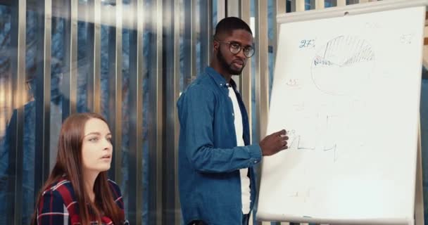 Un joven afroamericano con gafas y camisa de mezclilla escribe información a bordo. Grupo de estudiantes se prepara para los exámenes. — Vídeos de Stock