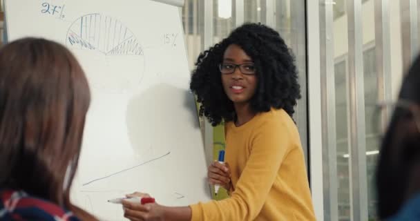 Joven chica afroamericana hermosa con gafas explica a grupo de estudiantes el tema de la conferencia y dibuja gráficos en pizarra. — Vídeos de Stock