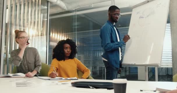 Jovem afro-americano com óculos em camisa jeans realiza levantamento entre estudantes do sexo feminino e escreve dados a bordo. — Vídeo de Stock