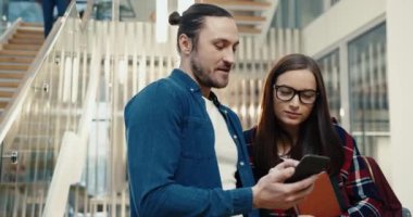 Kütüphanede dikilmiş sohbet eden iki genç öğrenci. Adam akıllı telefondan kızın komik videosunu gösteriyor..