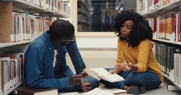 Twee jonge Afro-Amerikaanse studenten zitten op de vloer in de bibliotheek, bereiden examens voor, lezen boeken en bespreken ideeën. — Stockvideo