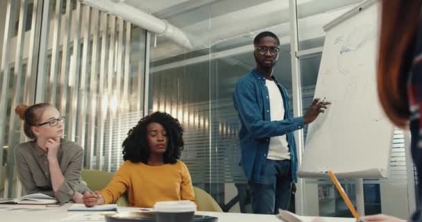 Los estudiantes se sientan en el aula y discuten la solución del problema. Joven afroamericano con gafas se para a bordo e ingresa datos. — Vídeo de stock