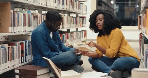 Två unga flitiga afroamerikanska studenter sitter på golvet i biblioteket och studerar litteratur noggrant inför tentorna. — Stockvideo