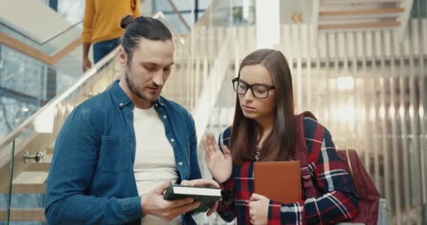 Twee jonge buitenlandse studenten communiceren in de universiteitslobby. Meisje vertelt verhaal, man is op zoek naar informatie over de telefoon. — Stockvideo